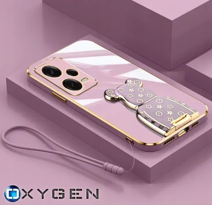 Na obrazie widoczne jest różowe etui na telefon z ozdobnym uchwytem w kształcie misia, ozdobionym gwiazdkami i kółkami. W lewym dolnym rogu znajduje się logo \'OXYGEN\'.