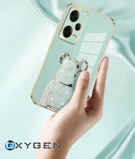 Osoba trzyma przezroczyste etui na telefon z ozdobnym motywem misia i złotymi krawędziami. W lewym dolnym rogu widoczne jest logo \'OXYGEN\'.
