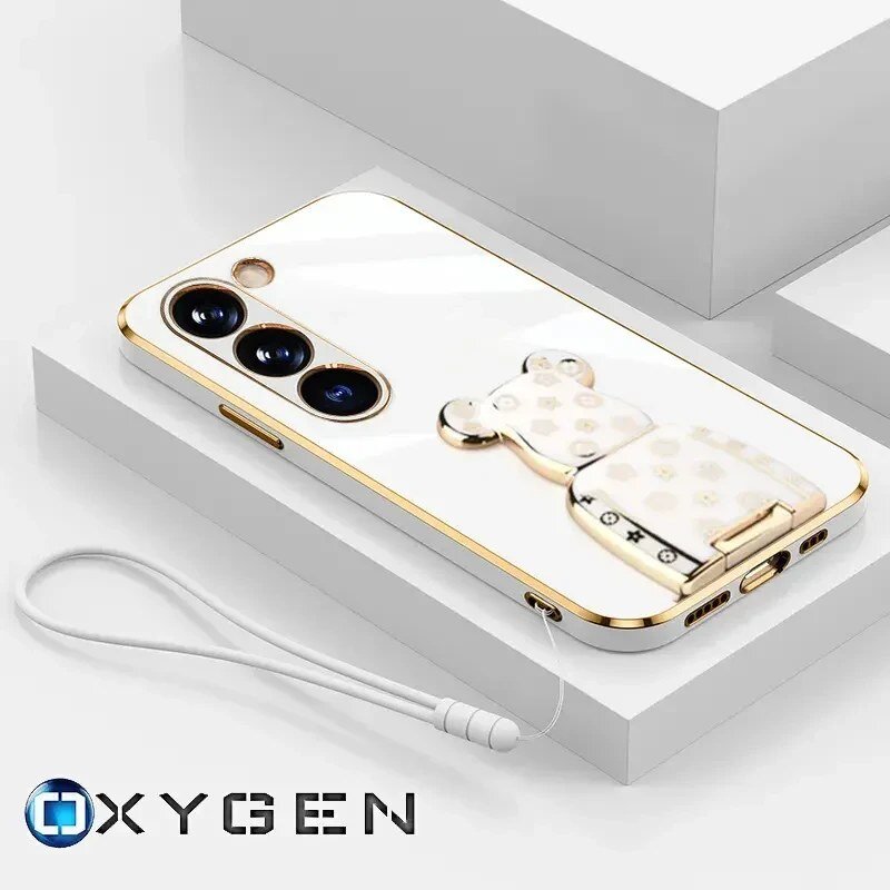 Na obrazie widoczne jest etui na telefon z trzema otworami na aparaty oraz ozdobnym uchwytem w kształcie misia. W dolnej części znajduje się logo \'OXYGEN\' oraz smycz przymocowana do etui.