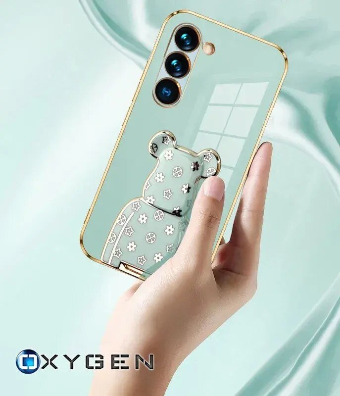 Osoba trzyma telefon w etui z przezroczystym tyłem i ozdobnym wzorem misia. Na dole widoczne jest logo \'OXYGEN\'.