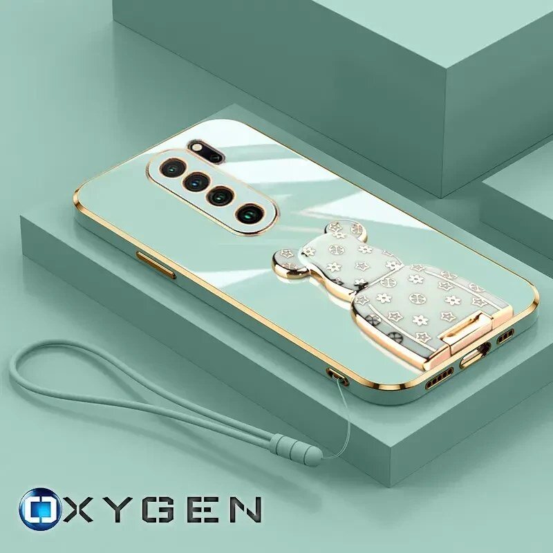 Na obrazie znajduje się etui na telefon z ozdobnym uchwytem w kształcie misia, umieszczone na tle w kolorze miętowym. Widoczne jest logo \'OXYGEN\' w dolnym rogu.