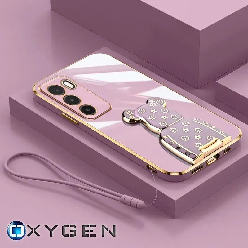 Różowe etui na telefon z metalicznymi elementami i uchwytem w kształcie misia, leży na różowym tle. Widoczne logo: OXYGEN.