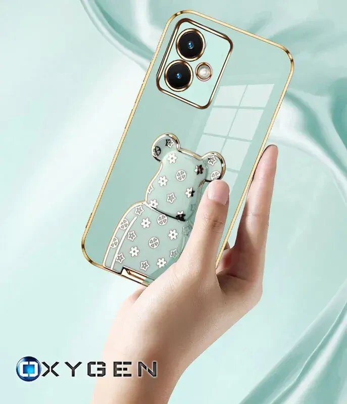 Osoba trzyma przezroczyste etui na telefon z ozdobnym wzorem misia i złotymi akcentami wokół aparatu. Na dole widoczne jest logo \'OXYGEN\'.