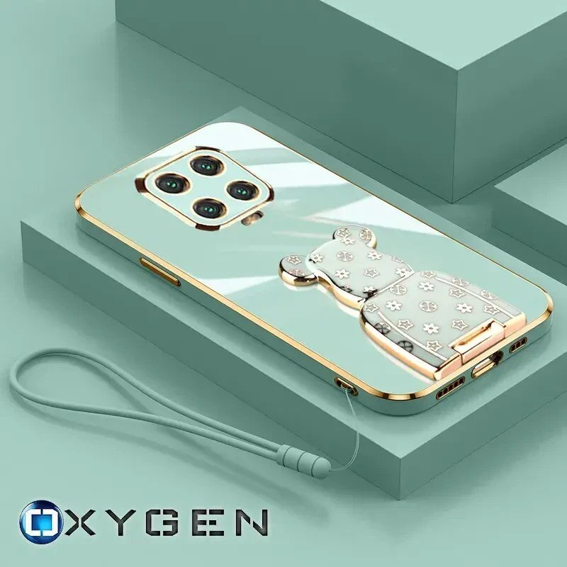 Na obrazie widoczne jest etui na telefon z metalicznym wykończeniem i ozdobnym uchwytem w kształcie misia. W dolnej części znajduje się logo \'OXYGEN\' oraz smycz przymocowana do etui.