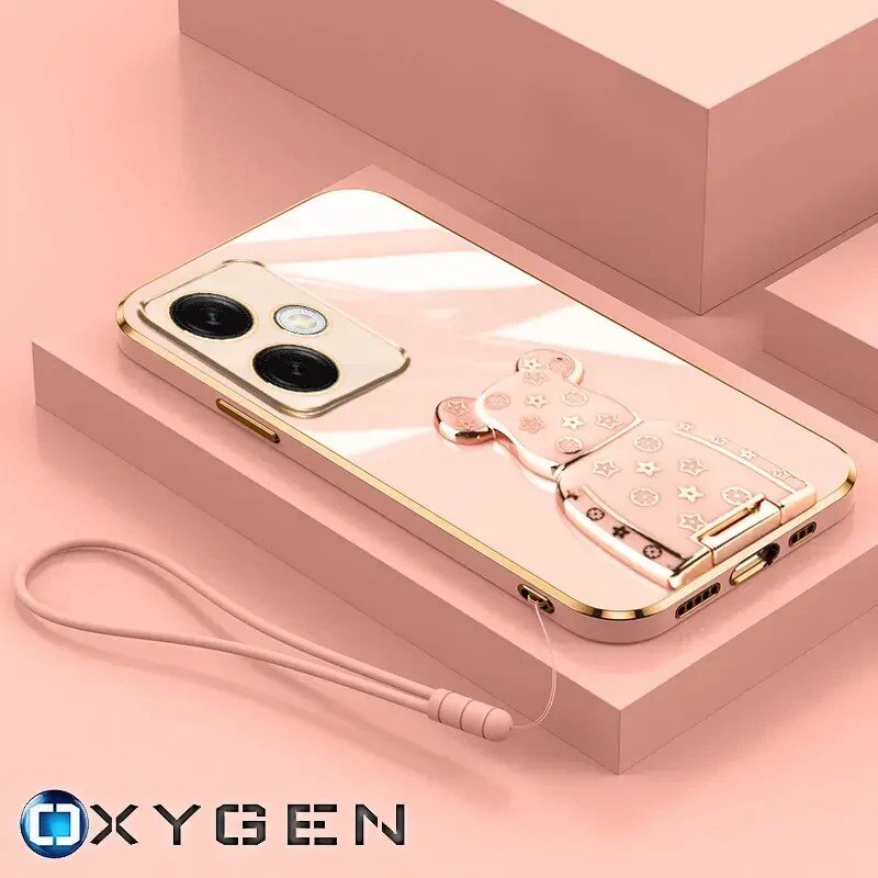 Na obrazie widoczne jest różowe etui na telefon z uchwytem w kształcie misia, leżące na różowej powierzchni. W dolnym rogu znajduje się logo \'OXYGEN\'.