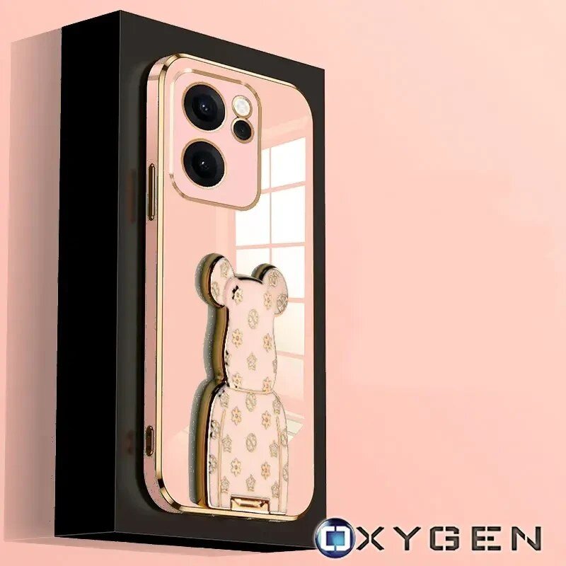 Etui na telefon w kolorze różowym z ozdobnym uchwytem w kształcie misia, umieszczone na czarnym tle. W prawym dolnym rogu widoczne jest logo \'OXYGEN\'.