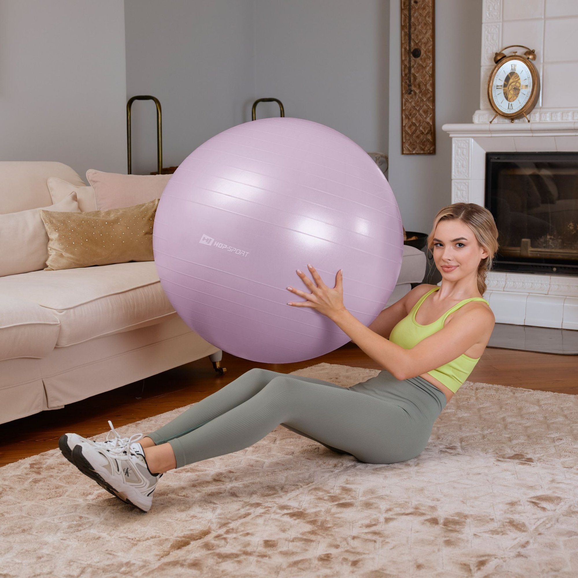 Osoba siedzi na dywanie w salonie, trzymając jasnofioletową piłkę fitness z logo \'HOP-SPORT\'. W tle widać sofę, kominek i zegar na półce.