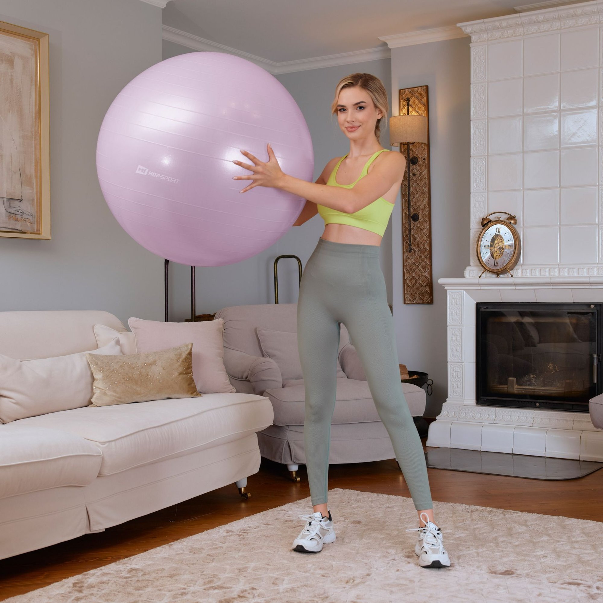 Osoba trzyma jasnofioletową piłkę fitness z logo \'HOP SPORT\' w salonie z kominkiem i sofą.