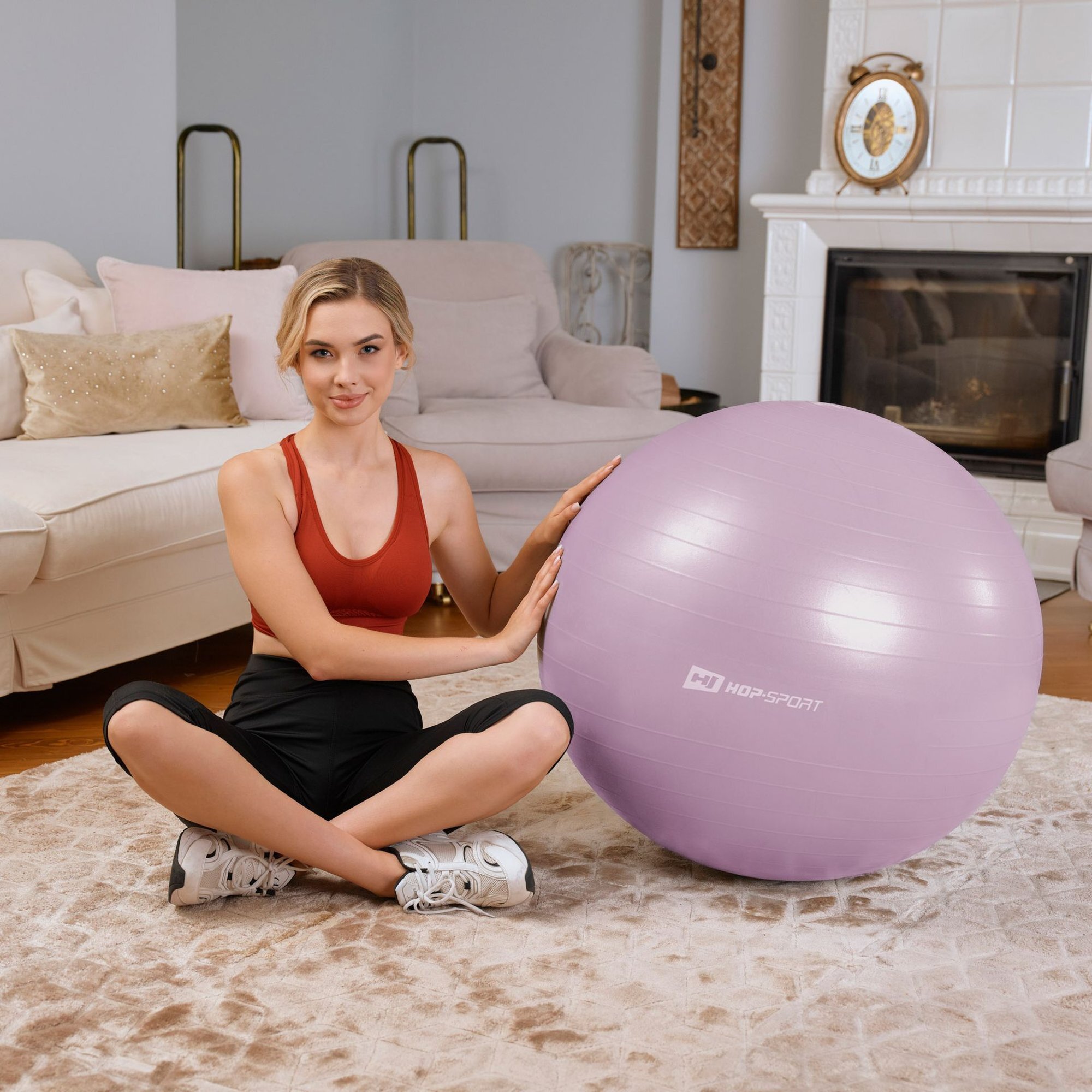 Osoba siedzi na dywanie obok jasnofioletowej piłki fitness z logo \'HOP-SPORT\'. W tle znajduje się jasna sofa i kominek.