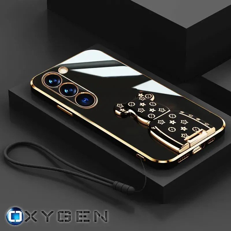 Czarne etui na telefon z trzema obiektywami aparatu i złotymi akcentami, posiada uchwyt w kształcie misia z wzorem gwiazdek. Widoczne jest logo OXYGEN oraz czarna smycz przymocowana do etui.