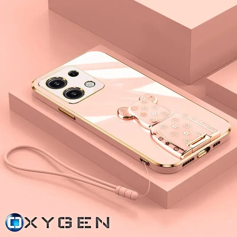 Na obrazie widoczne jest różowe etui na telefon z uchwytem w kształcie misia, leżące na różowej powierzchni. W dolnym rogu znajduje się logo \'OXYGEN\'.