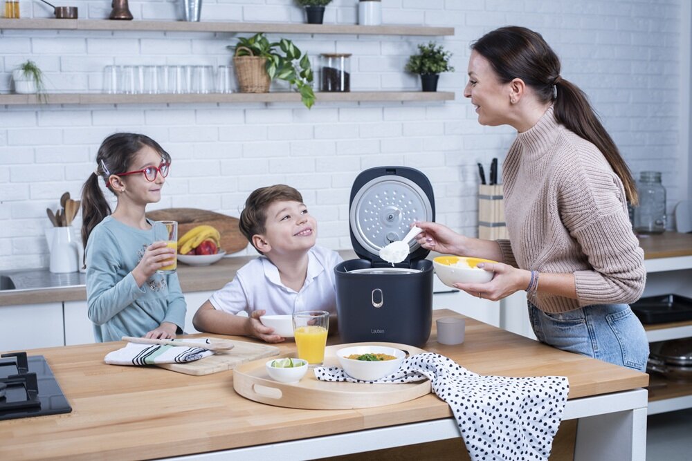 Kobieta używa ryżowaru marki Lauben w kuchni, podczas gdy dwoje dzieci siedzi przy stole z napojami i jedzeniem. Na blacie znajdują się miski z jedzeniem, szklanki z sokiem i ręcznik kuchenny w kropki.