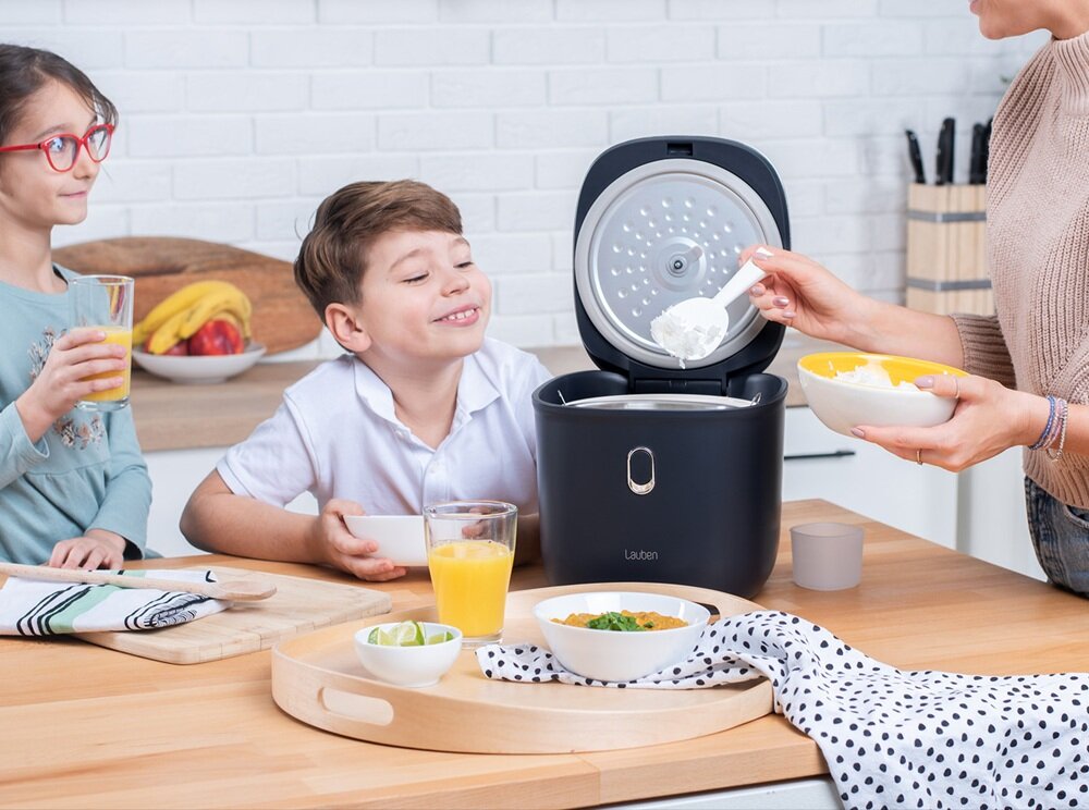 Na blacie kuchennym stoi ryżowar marki Lauben z otwartą pokrywą, a obok znajdują się miski z jedzeniem i szklanki z sokiem. W tle widać białą ceglaną ścianę i drewnianą deskę z owocami.