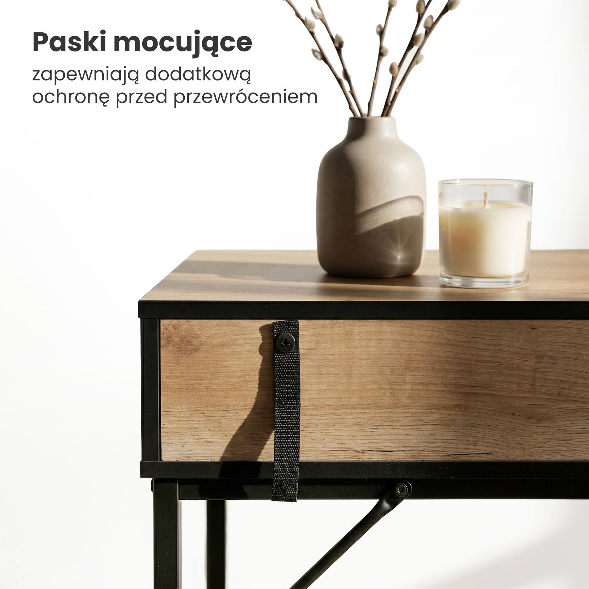 Na drewnianym blacie stolika znajduje się szara waza z gałązkami oraz zapalona świeca w szklanym pojemniku. Obok widoczny jest czarny pasek mocujący na froncie szuflady z tekstem: \'Paski mocujące zapewniają dodatkową ochronę przed przewróceniem\'.