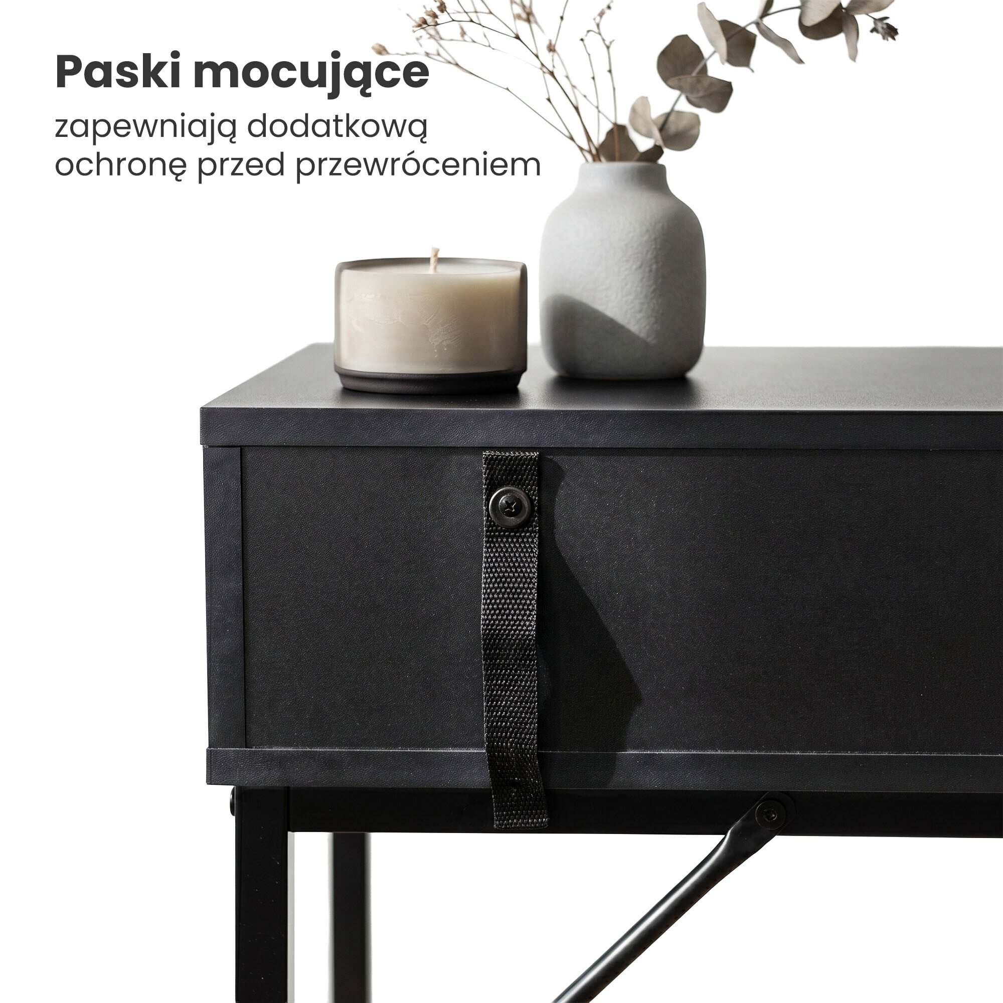 Czarny stolik z szufladą, na którym stoją świeca i wazon z suchymi gałązkami. Tekst na obrazie: \'Paski mocujące zapewniają dodatkową ochronę przed przewróceniem\'.