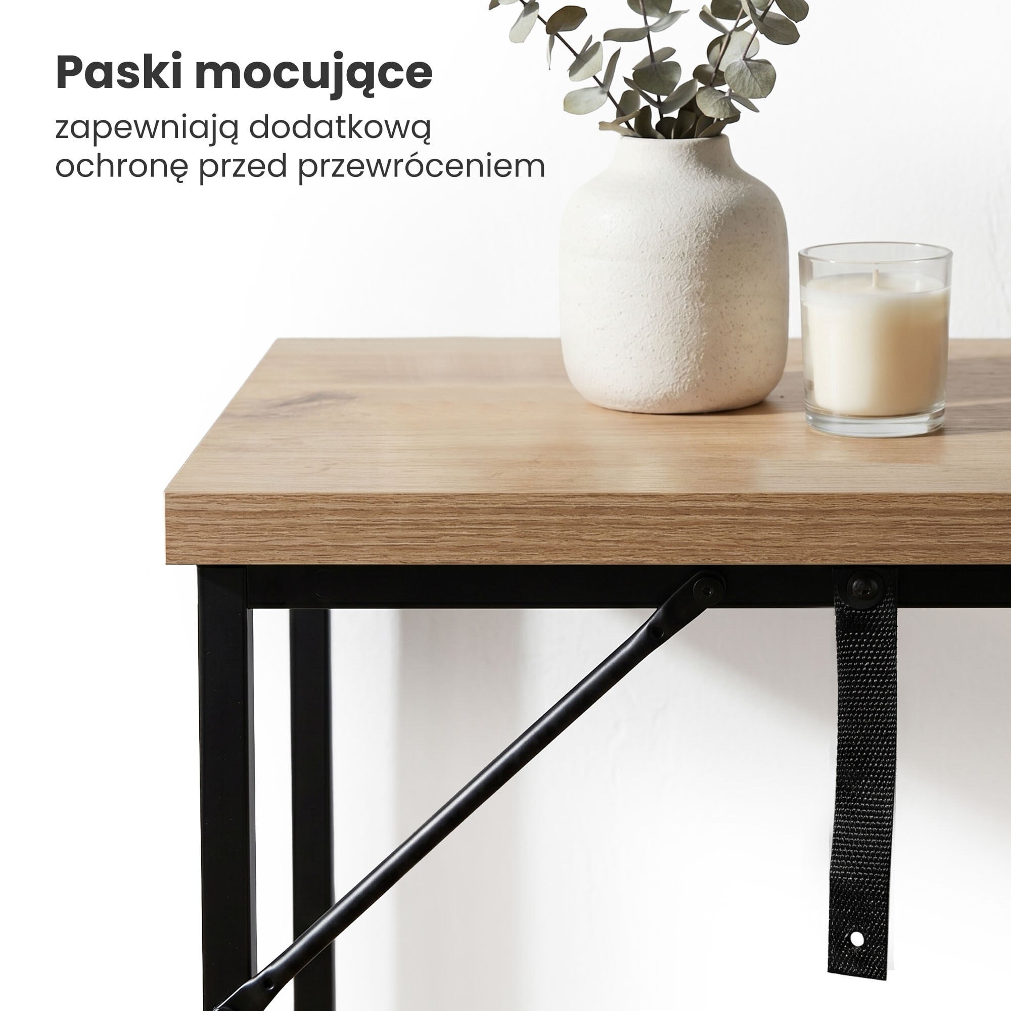 Na drewnianym blacie stolika znajduje się biała wazon z rośliną oraz świeca w szklanym naczyniu. Obok widoczny jest czarny pasek mocujący, a w tle tekst: \'Paski mocujące zapewniają dodatkową ochronę przed przewróceniem\'.