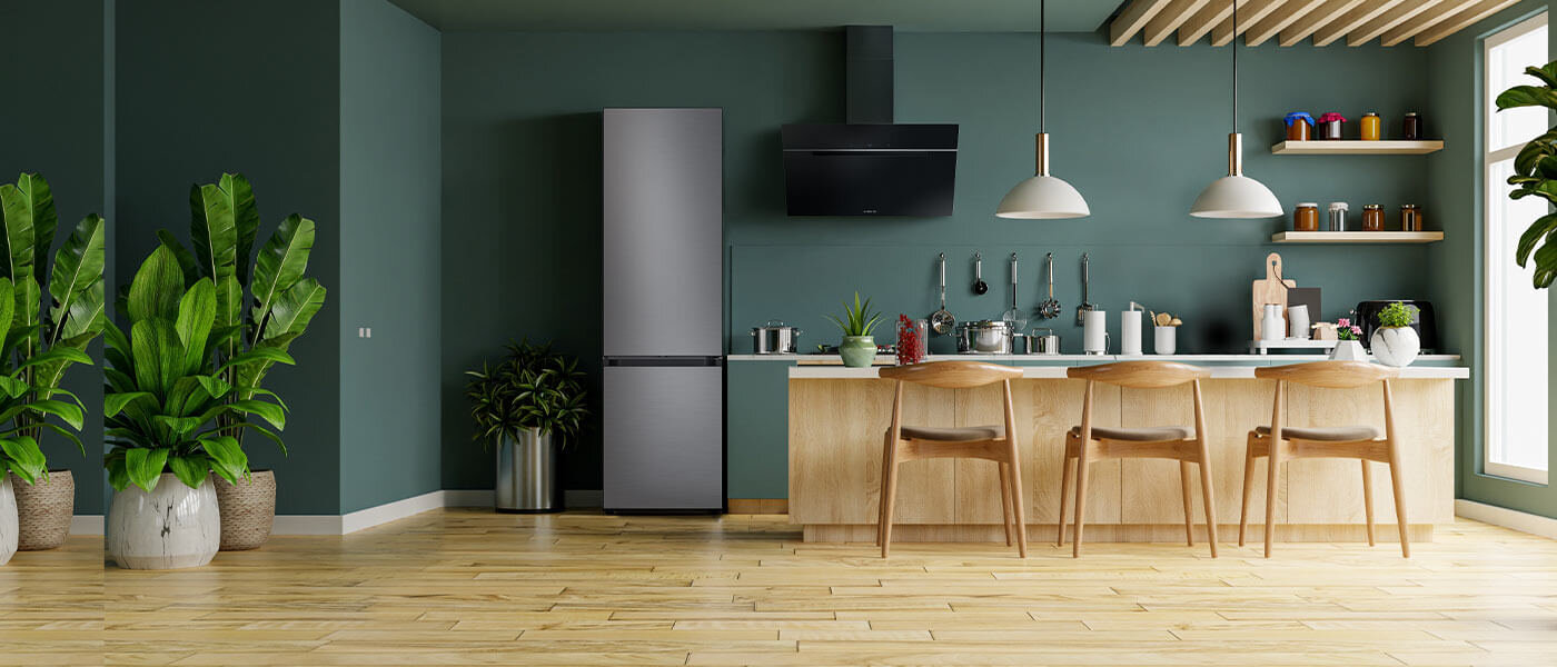 Ciemnozielone wnętrze kuchenne z dwudrzwiową lodówką Samsung RB38C7B5DS9 z oferty sklepu Media Expert Kuchnia z drewnianą podłogą, w której znajduje się lodówka w kolorze szczotkowanej stali nierdzewnej. Obok lodówki jest wyspa kuchenna z krzesłami, a na blacie stoją różne przedmioty kuchenne.