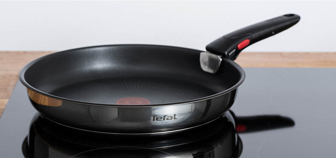 Na obrazie widoczna jest czarna patelnia z metalicznym wykończeniem, umieszczona na płycie indukcyjnej. Na boku patelni znajduje się logo \'Tefal\'.