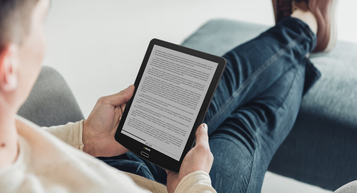 Osoba trzyma czarny czytnik e-booków z wyświetlonym tekstem na ekranie. Urządzenie jest używane w pozycji siedzącej na sofie.