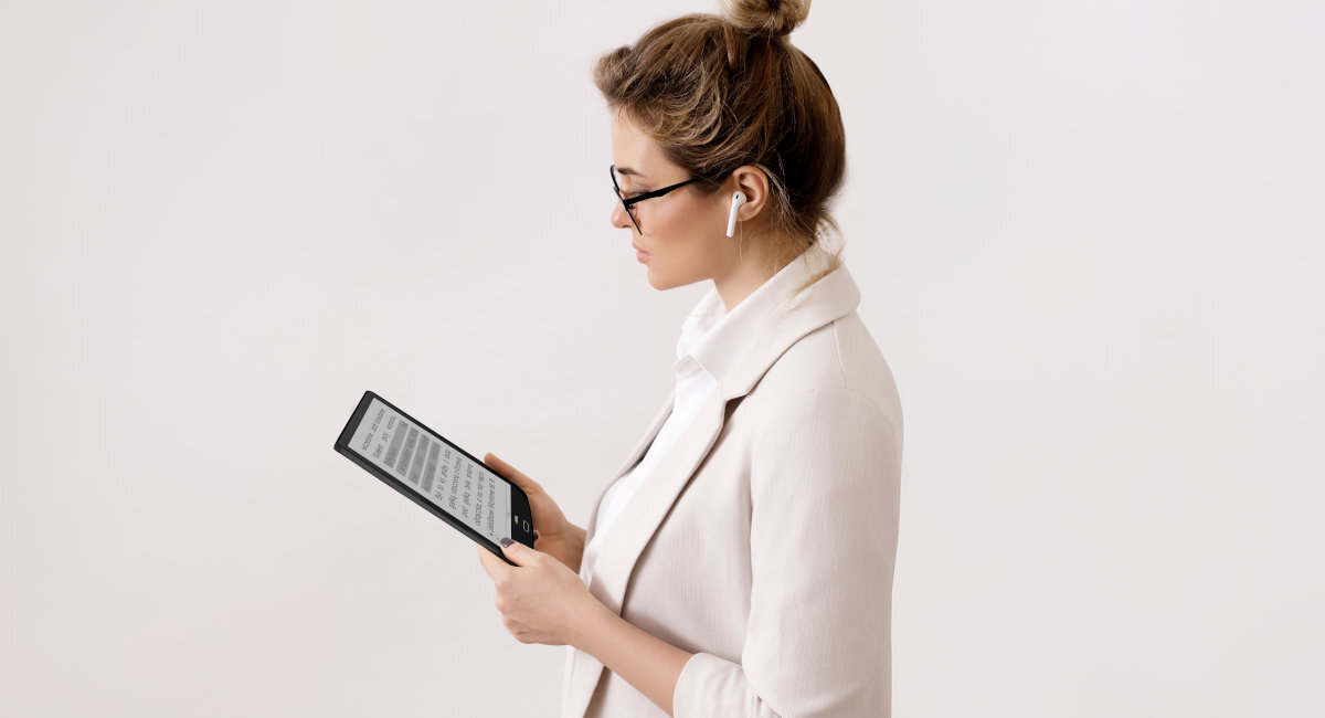 Osoba trzymająca czytnik e-booków z widocznym tekstem na ekranie. Osoba ma na sobie jasne ubranie i nosi okulary oraz słuchawki bezprzewodowe.