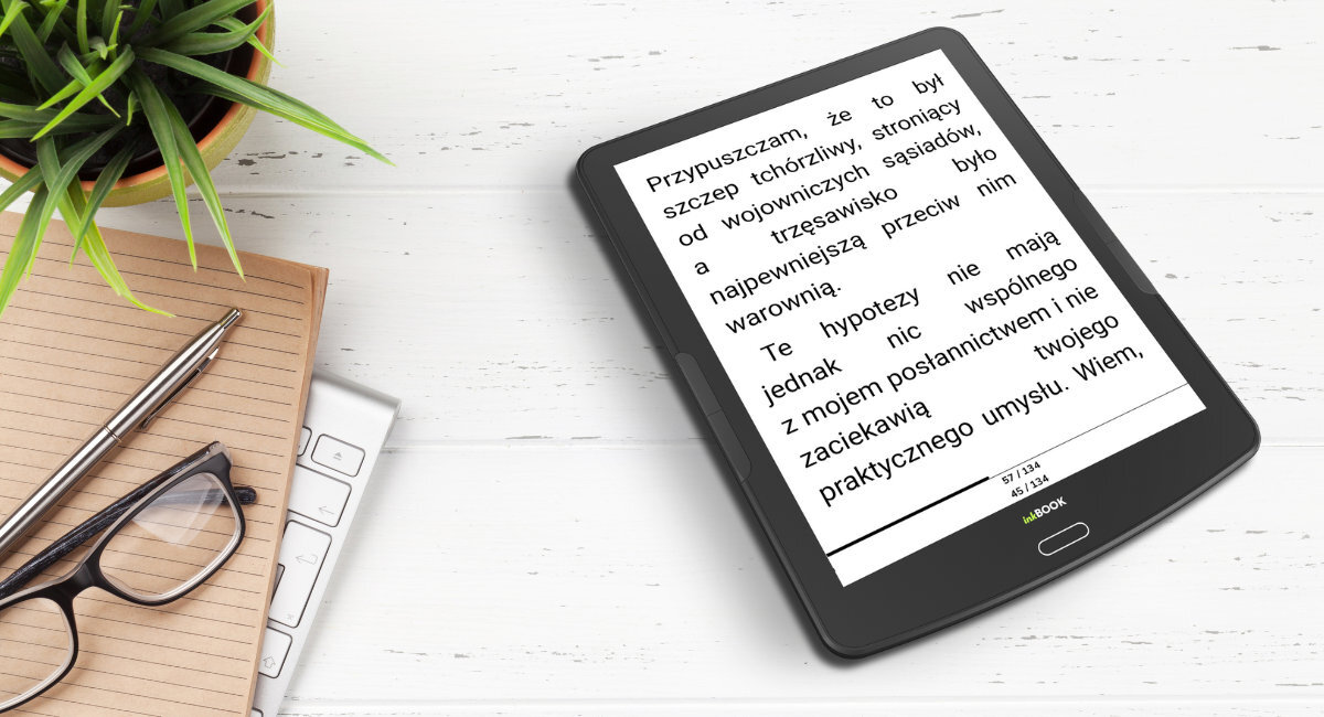 Na białym drewnianym tle leży czarny czytnik e-booków z wyświetlonym tekstem: \'Przypuszczam, że to był szczep tchórzliwy, stroniący od wojowniczych sąsiadów, a trzęsawisko było najpewniejszą przeciw nim warownią. Te hipotezy nie mają jednak nic wspólnego z mojem posłannictwem i nie zaciekawią twojego praktycznego umysłu. Wiem.\'. Obok znajduje się notatnik, okulary, długopis i klawiatura, a w lewym górnym rogu widoczna jest zielona roślina.
