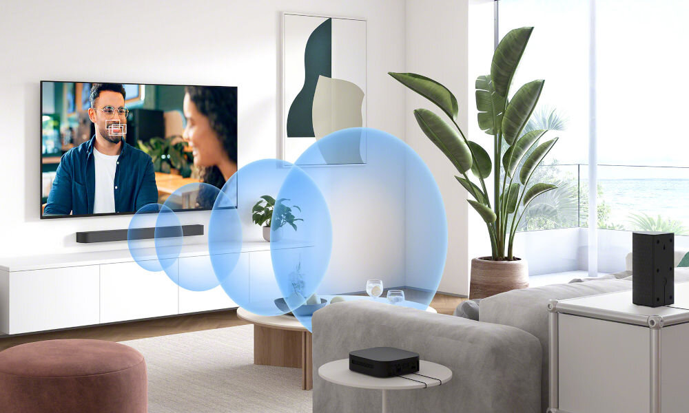 W nowoczesnym salonie znajduje się telewizor z włączonym programem oraz soundbar umieszczony pod nim. W pomieszczeniu widoczne są również rośliny doniczkowe i nowoczesne meble.