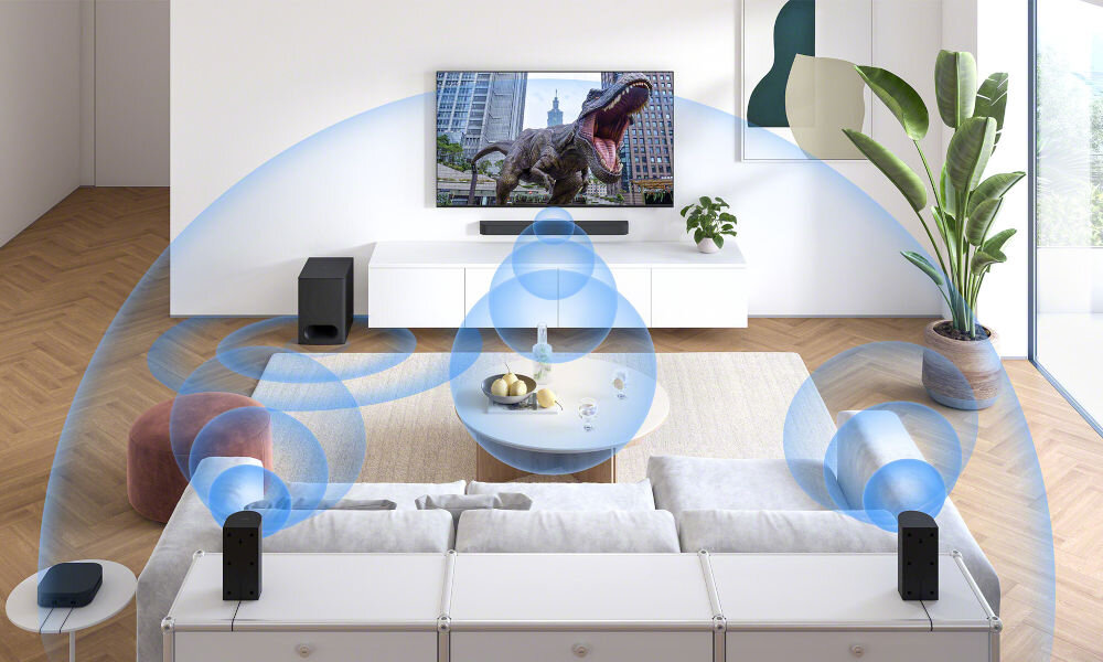 W salonie znajduje się telewizor z wyświetlonym obrazem dinozaura, a przed nim soundbar i subwoofer. Wokół pomieszczenia widoczne są graficzne fale dźwiękowe, sugerujące przestrzenny dźwięk.