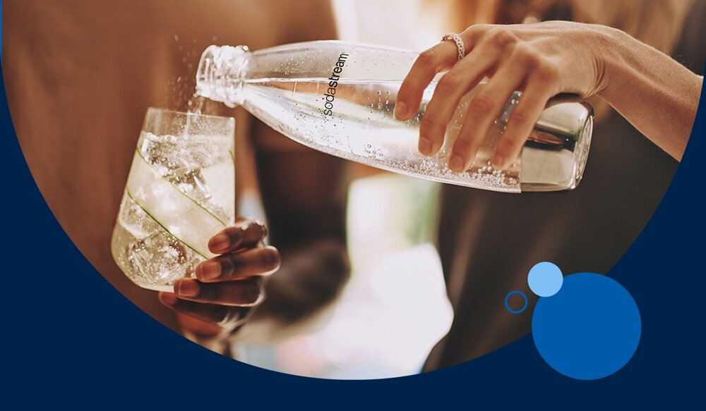 Osoba nalewa wodę z butelki z napisem \'sodastream\' do szklanki z lodem i plasterkami limonki. W tle widoczne są rozmyte kształty i kolory.