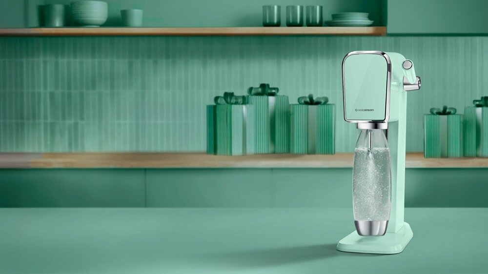 Miętowy saturator stoi na blacie w kuchni, w tle widać półkę z naczyniami i dekoracjami w odcieniach zieleni. Na urządzeniu widoczne jest logo \'SODASTREAM\'.