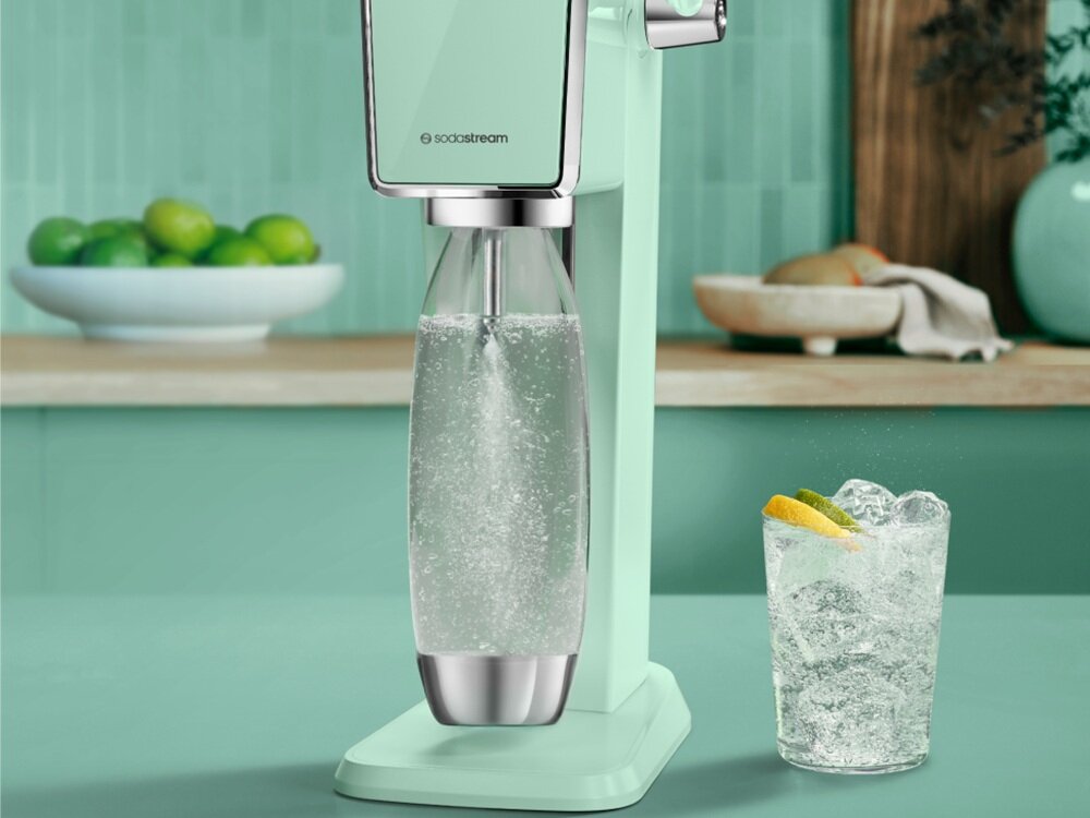 Na obrazie widoczny jest miętowy saturator z logo \'sodastream\', z przymocowaną butelką wypełnioną gazowaną wodą. W tle znajduje się szklanka z napojem i plasterkami cytryny oraz miska z limonkami.