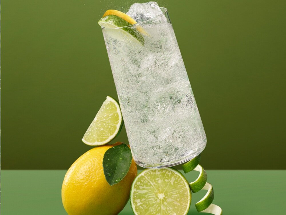 Szklanka z napojem gazowanym i plasterkiem cytryny oraz limonki, otoczona kawałkami cytrusów na zielonym tle.