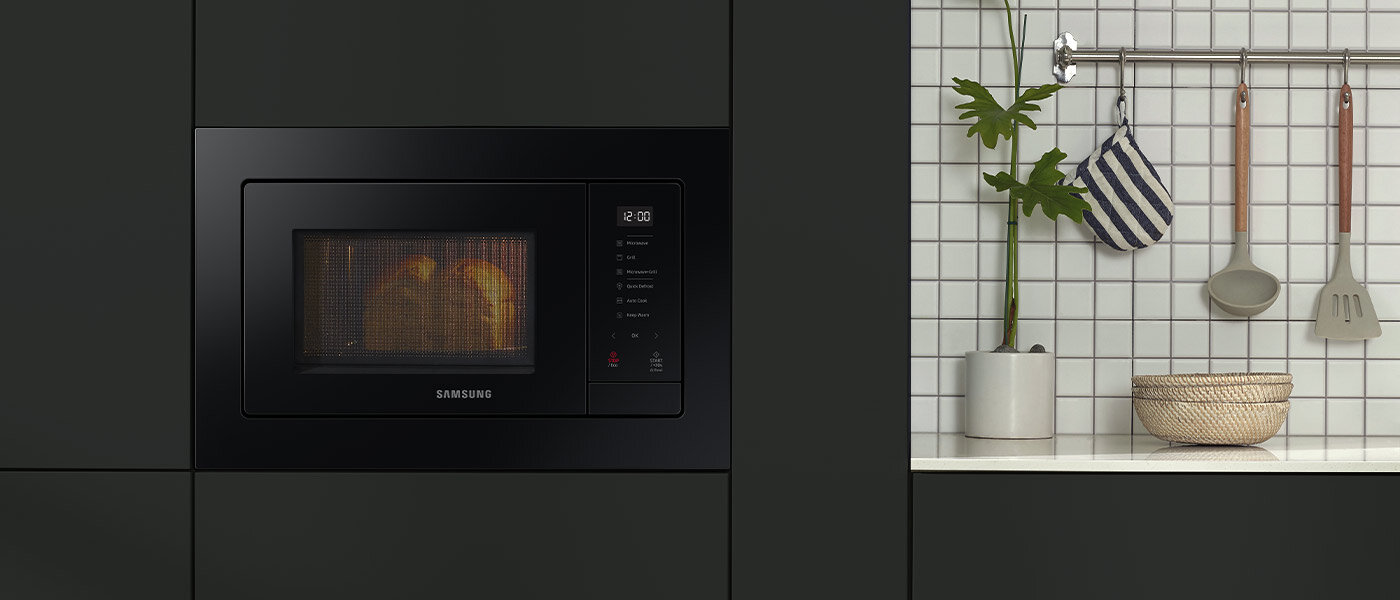 Elegancka i funkcjonalna kuchenka mikrofalowa Samsung Czarna kuchenka mikrofalowa wbudowana w szafkę, z widocznym logo Samsung na drzwiczkach. Obok znajduje się biała ściana z kafelkami, na której wiszą przybory kuchenne i roślina w doniczce.