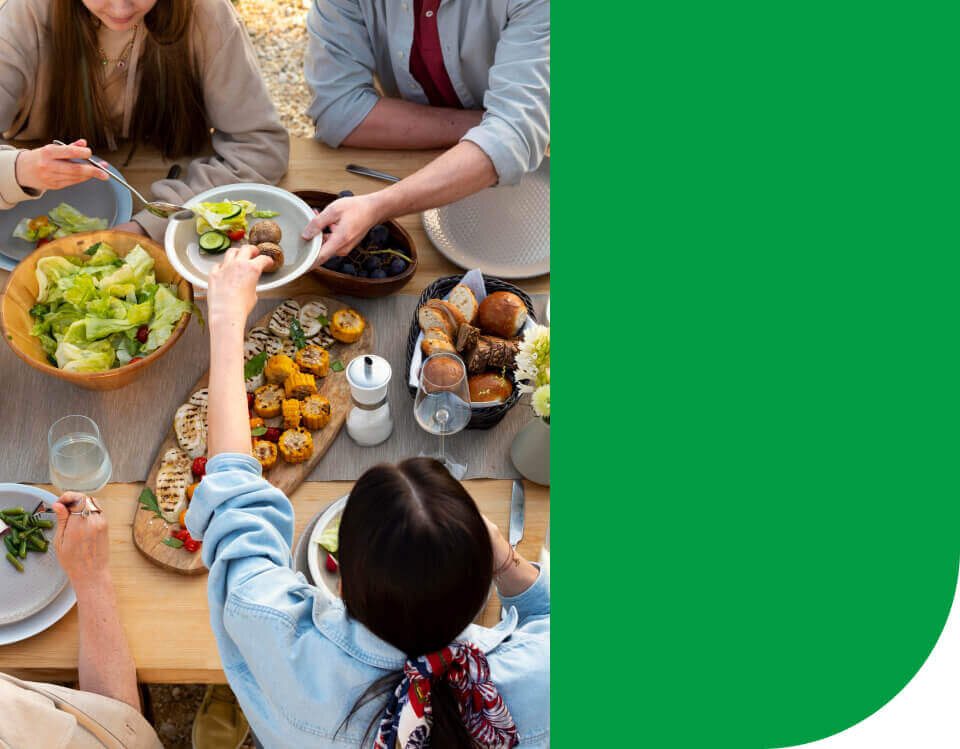 Grupa osób siedzi przy stole zastawionym różnorodnymi potrawami, w tym sałatką, pieczywem i grillowanymi warzywami. Obok stołu znajduje się zielony panel.
