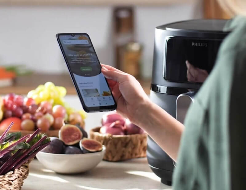 Osoba trzyma smartfon z otwartą aplikacją, obok znajduje się frytkownica beztłuszczowa z widocznym logo PHILIPS. W tle widać owoce i warzywa na blacie kuchennym.