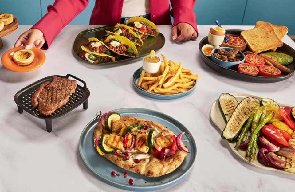 Na stole znajduje się różnorodność potraw, w tym tacos, frytki, grillowane warzywa, stek oraz pieczywo z dodatkami. Osoba w czerwonej marynarce trzyma talerz z małą tartą.