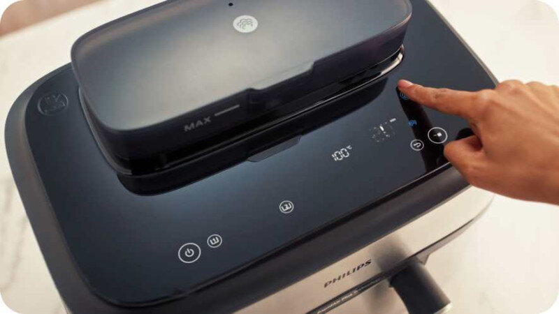 Osoba dotyka panelu dotykowego frytkownicy beztłuszczowej z wyświetlaczem pokazującym temperaturę 100°C. Na panelu widoczne są ikony i logo PHILIPS.