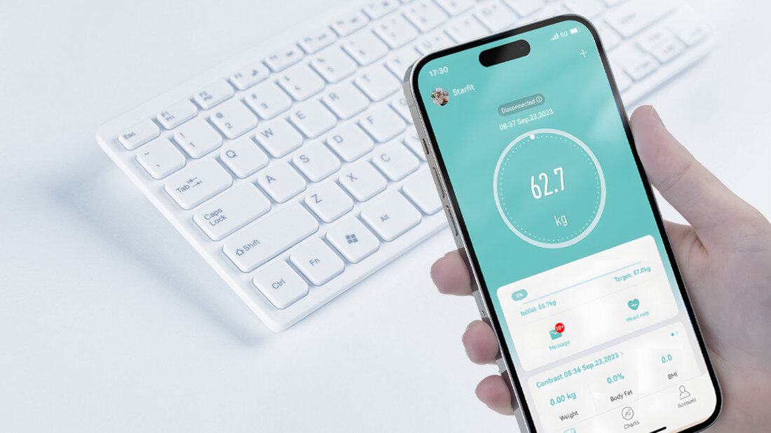 Osoba trzyma smartfon z aplikacją pokazującą wagę 62.7 kg. W tle widoczna jest biała klawiatura.