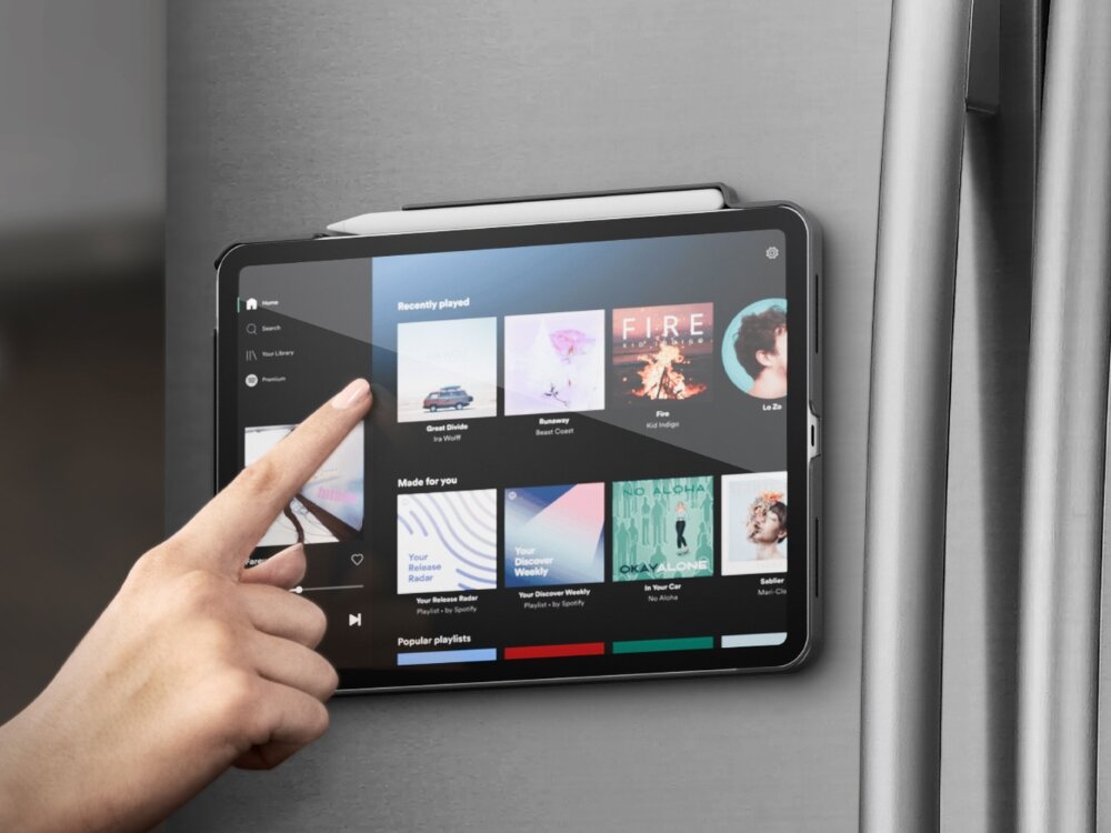 Tablet przymocowany do metalowej powierzchni, na ekranie widoczna jest aplikacja muzyczna z listą odtwarzania. Dłoń wskazuje na ekran, wybierając jedną z opcji.