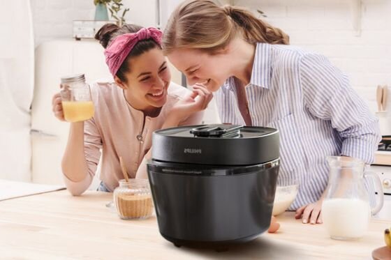 Na blacie kuchennym stoi czarny multicooker z widocznym logo PHILIPS. W tle dwie osoby rozmawiają i trzymają szklanki z napojami.