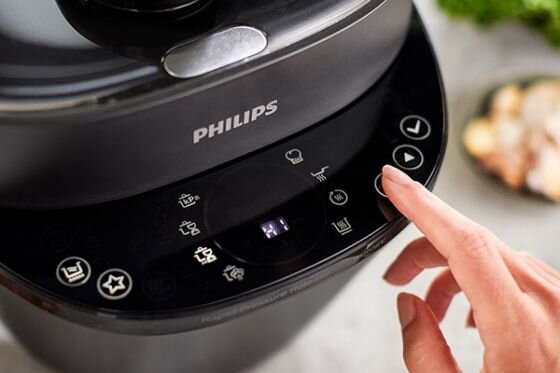 Czarna płyta sterująca multicookera z widocznymi ikonami funkcji i przyciskami dotykowymi. Logo \'PHILIPS\' znajduje się na górze urządzenia, a dłoń naciska jeden z przycisków.