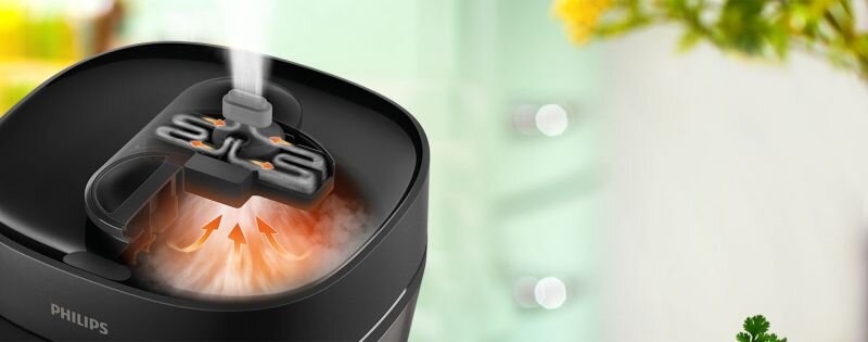 Czarny multicooker z widocznym logo PHILIPS, z którego unosi się para przez otwór w pokrywie. W tle rozmyte elementy kuchenne i rośliny.