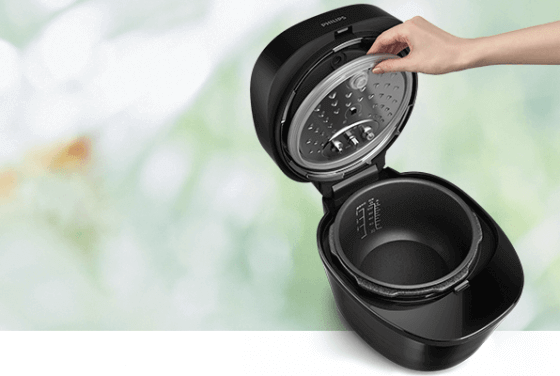 Na obrazie widoczny jest czarny multicooker z otwartą pokrywą, trzymaną przez rękę. Wnętrze urządzenia jest puste, a na pokrywie widnieje logo \'PHILIPS\'.