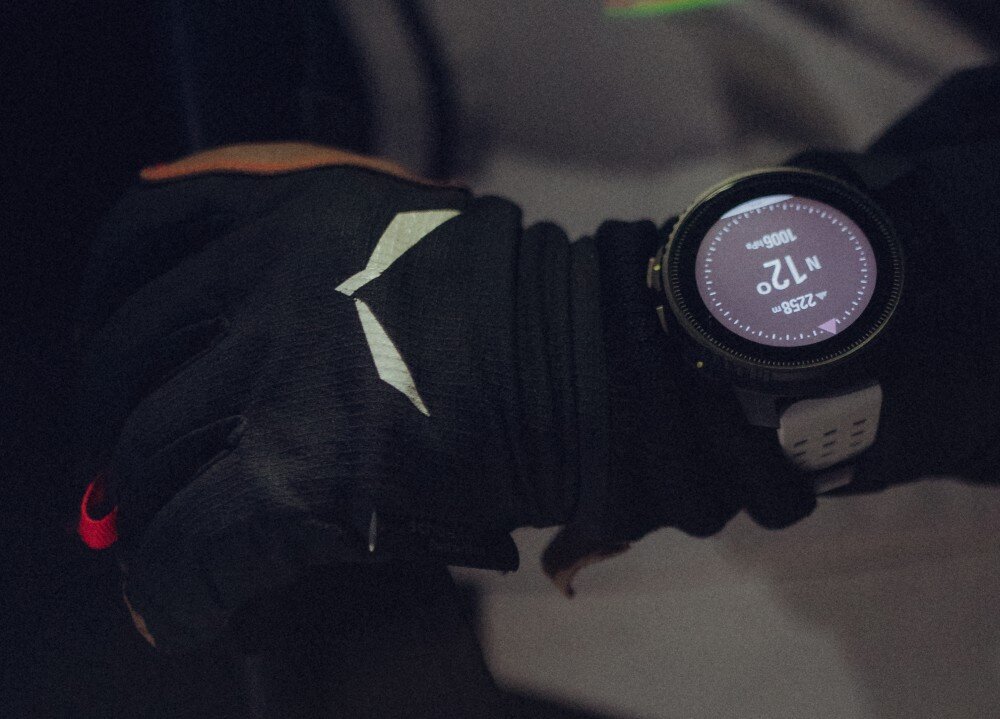 Osoba nosi czarne rękawiczki i smartwatch z okrągłym wyświetlaczem, na którym widoczny jest kompas z kierunkiem \'N\' oraz liczba \'12\'.