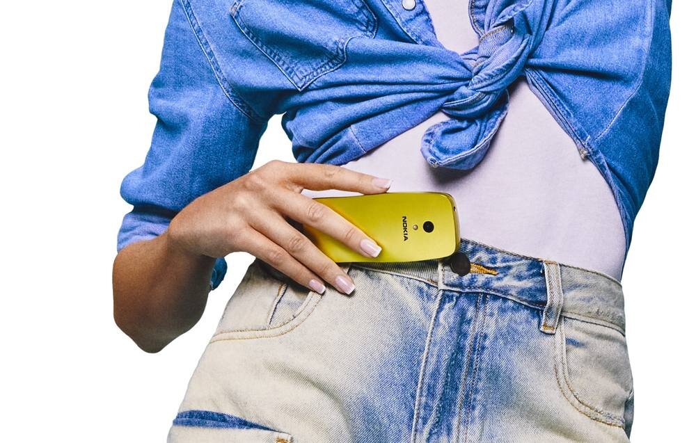 Osoba trzyma złoty telefon z napisem \'NOKIA\' wkładając go do kieszeni spodni. Osoba ma na sobie niebieską koszulę zawiązaną na supeł.