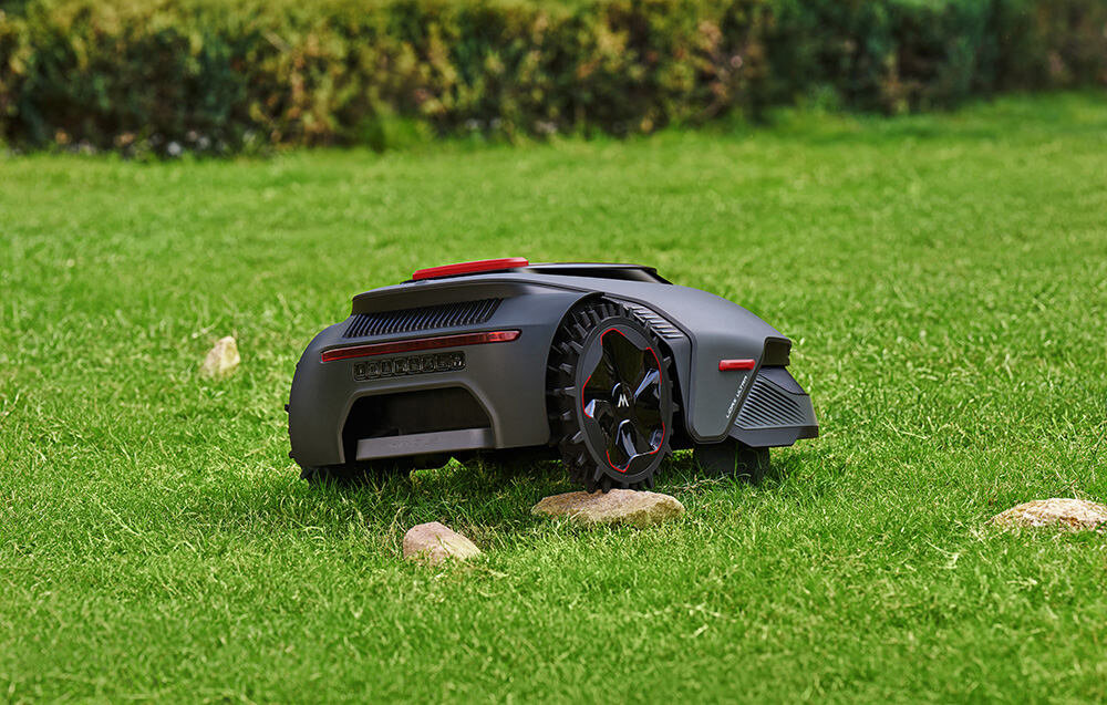 Na obrazie widoczny jest robot koszący o nowoczesnym designie, poruszający się po trawniku. W tle znajdują się krzewy, a na trawie leżą kamienie.