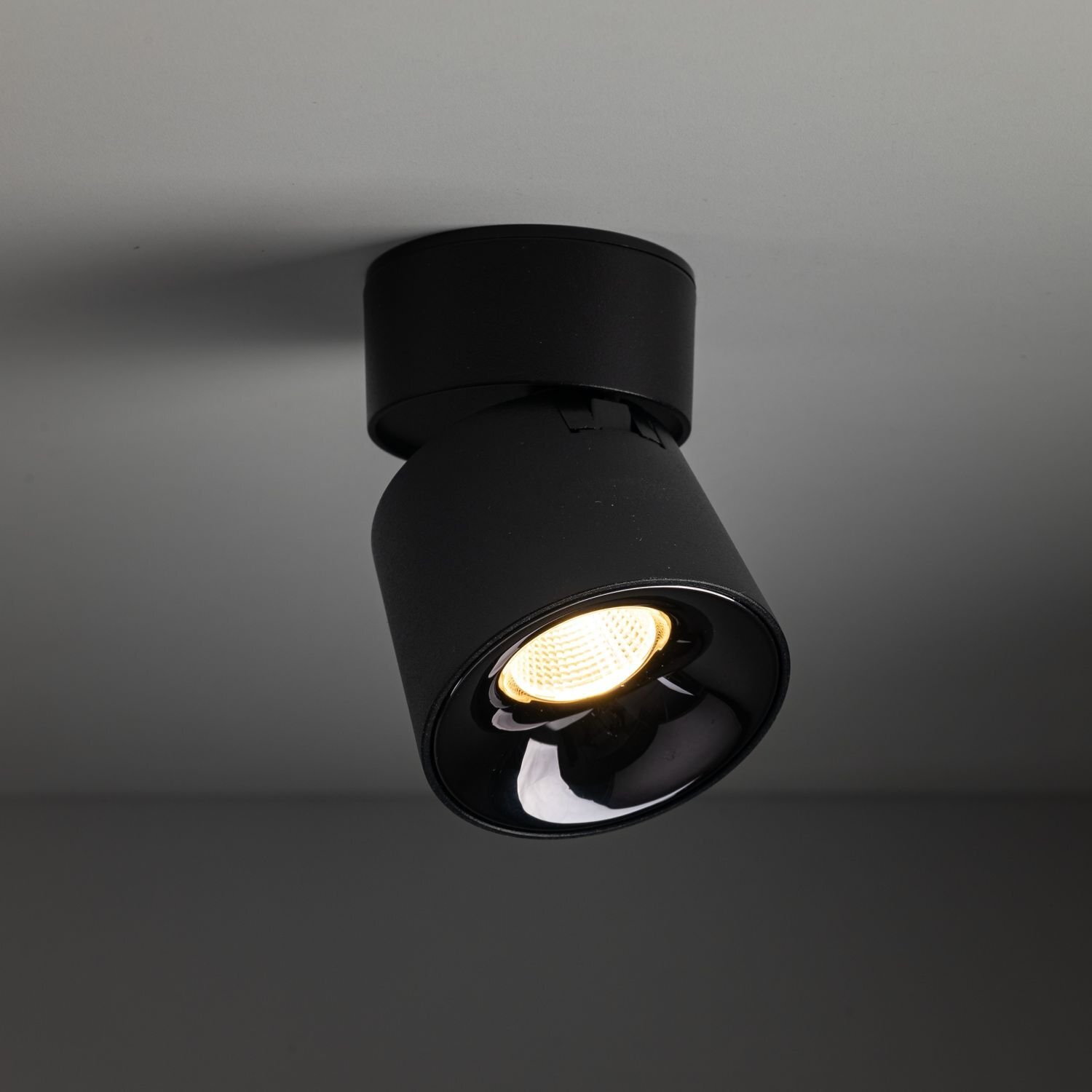 Czarna lampa reflektorowa LED zamontowana na suficie, emitująca ciepłe białe światło. Lampa ma cylindryczny kształt i jest ruchoma.