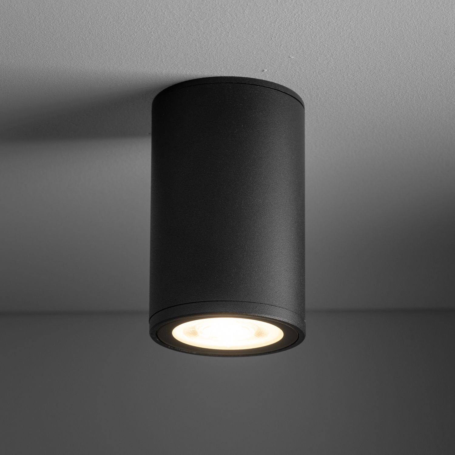 Czarna, cylindryczna oprawa natynkowa z metalu, emitująca światło w dół. Zamontowana na suficie, ma nowoczesny design.