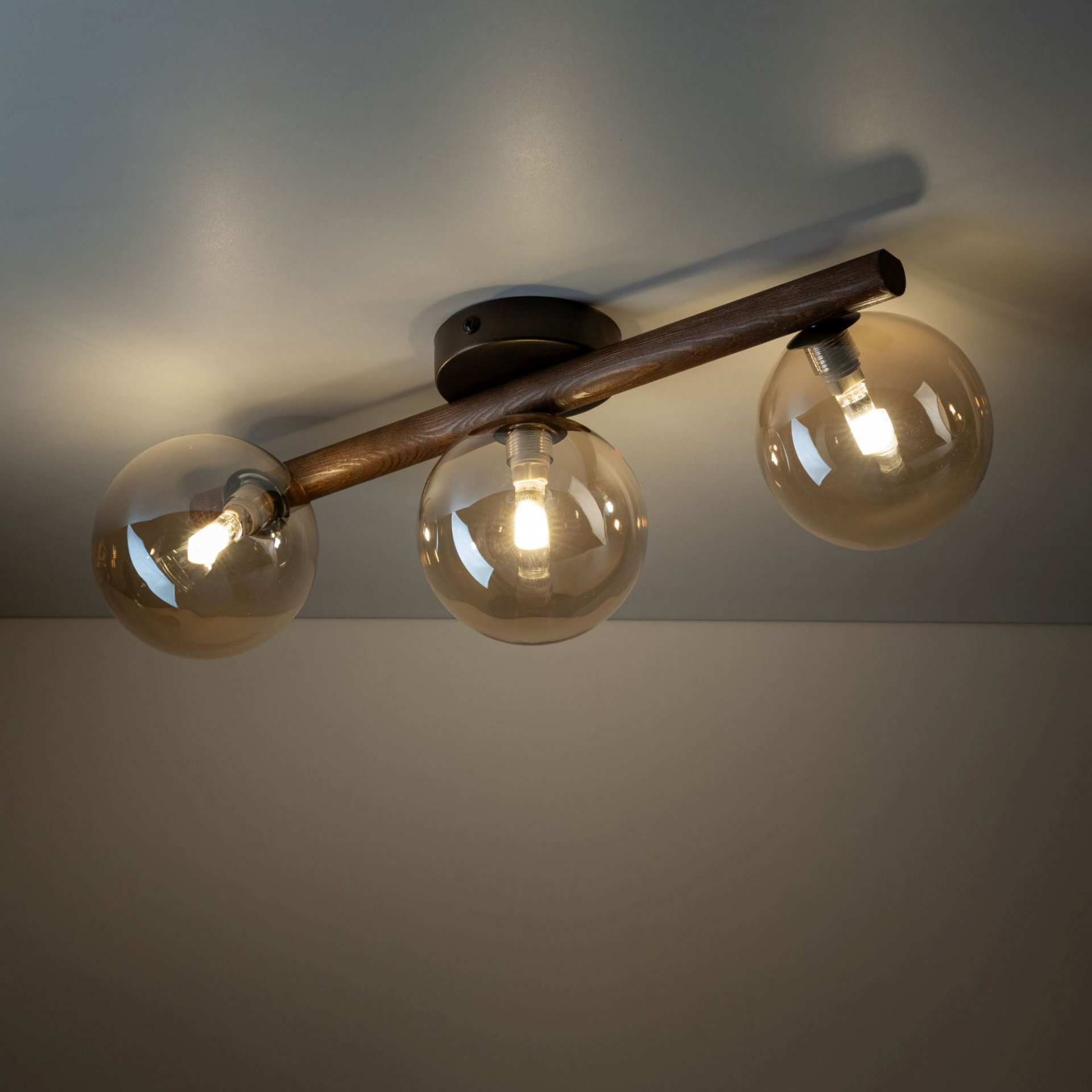 Lampa sufitowa z trzema bursztynowymi kloszami zamontowanymi na drewnianej belce. Każdy klosz zawiera widoczną żarówkę, a całość przymocowana jest do sufitu za pomocą ciemnej podstawy.