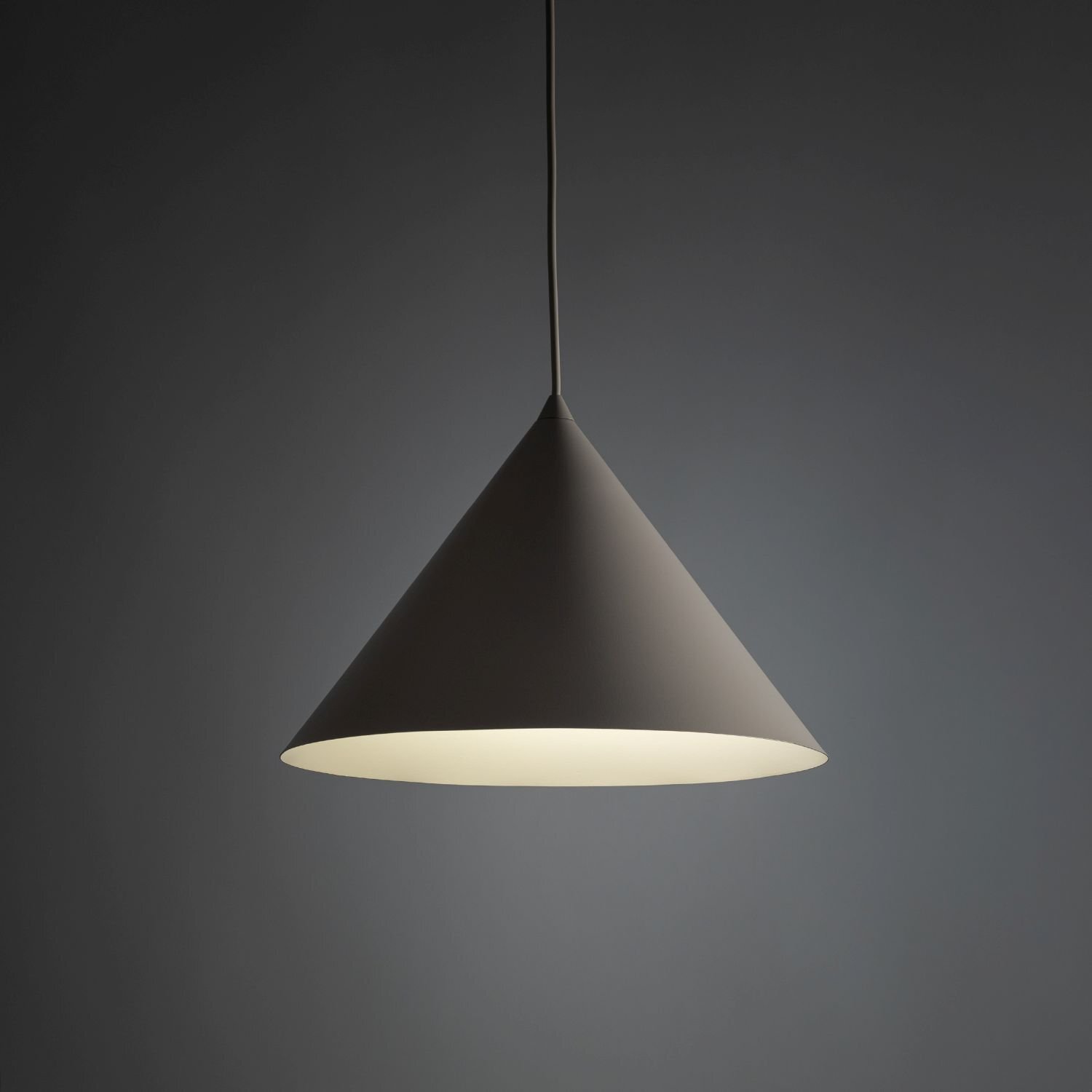 Na obrazie widoczna jest nowoczesna lampa wisząca w kształcie stożka, w kolorze kaszmiru, z jasnym wnętrzem. Lampa jest zawieszona na cienkim przewodzie na tle ciemnoszarej ściany.