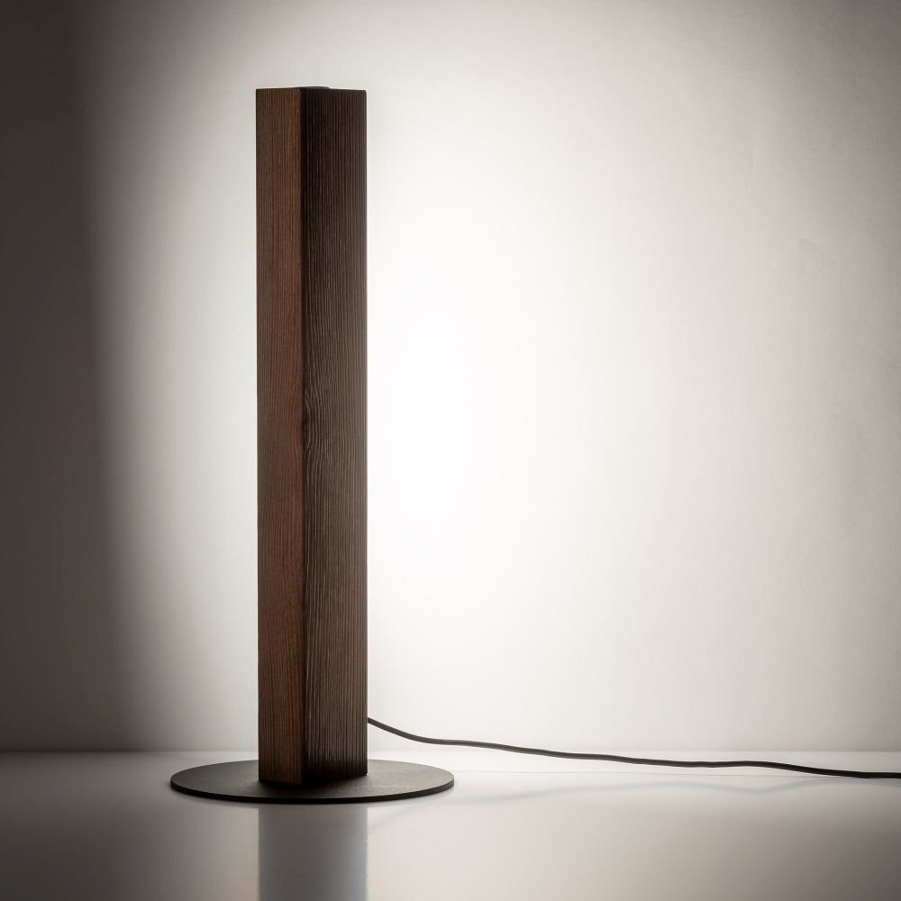 Na obrazie widoczna jest nowoczesna lampa stołowa o prostokątnym, drewnianym korpusie w kolorze orzecha, umieszczona na okrągłej, czarnej podstawie. Lampa emituje ciepłe, białe światło, a z tyłu widoczny jest przewód zasilający.
